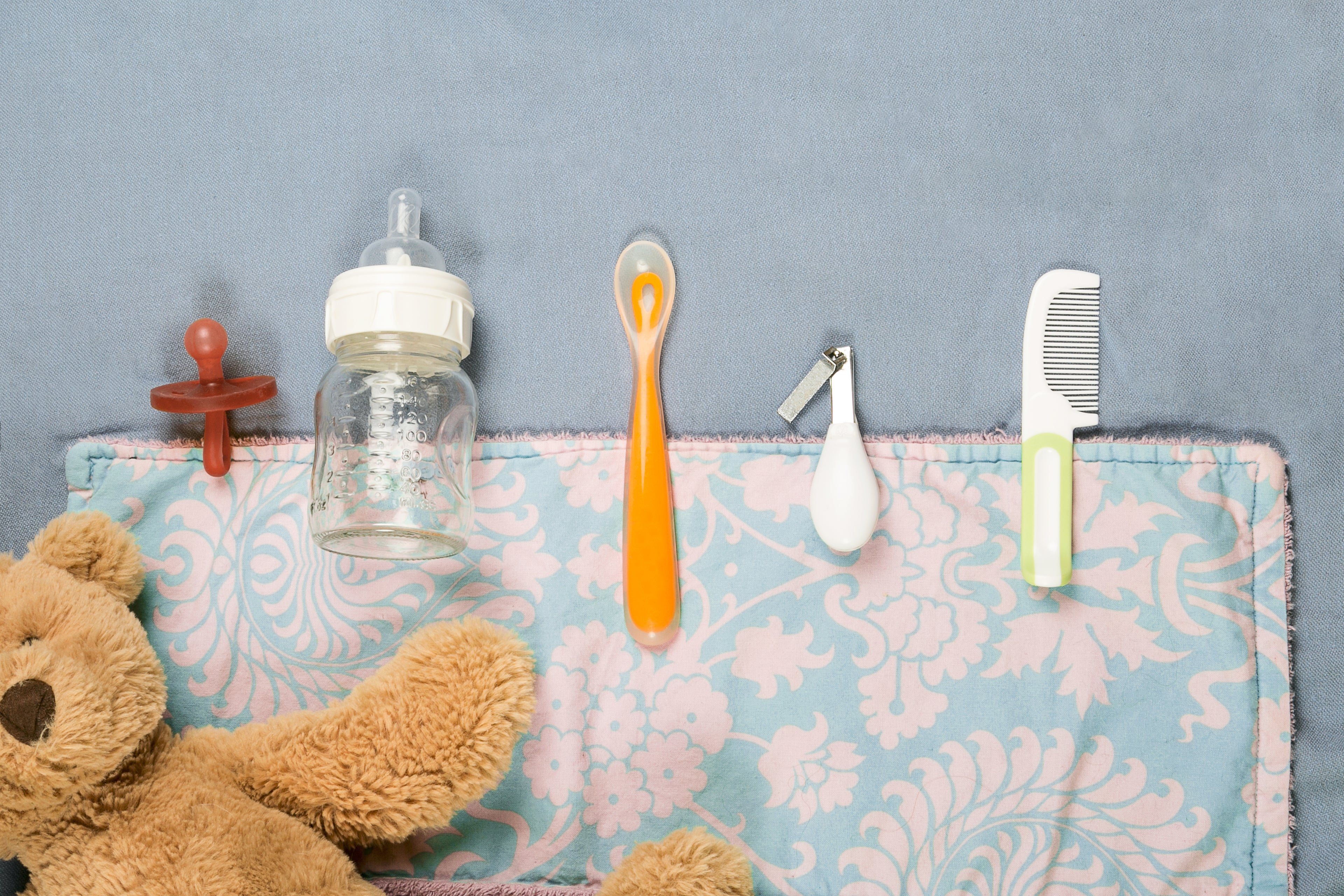 Baby items including a bottle, spoon, and teddy bear on a floral blanket against a blue wall.